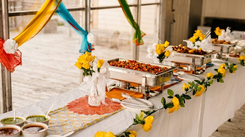 Vibrant Indian buffet table with chafing dishes, sauces, and yellow floral decorations.