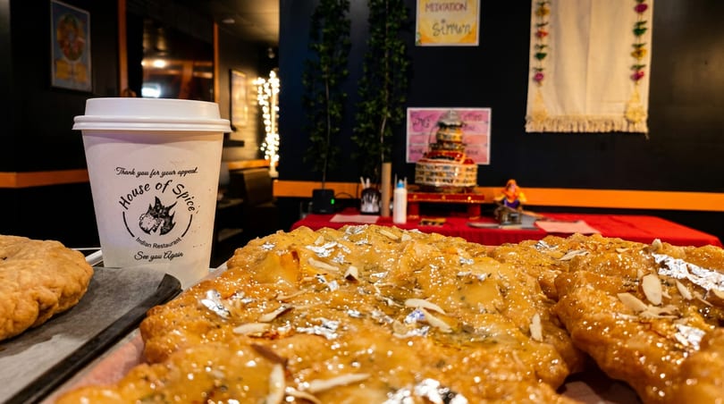 Glazed Indian sweets with almonds and silver leaf, beside a 'House of Spice' cup in a restaurant.