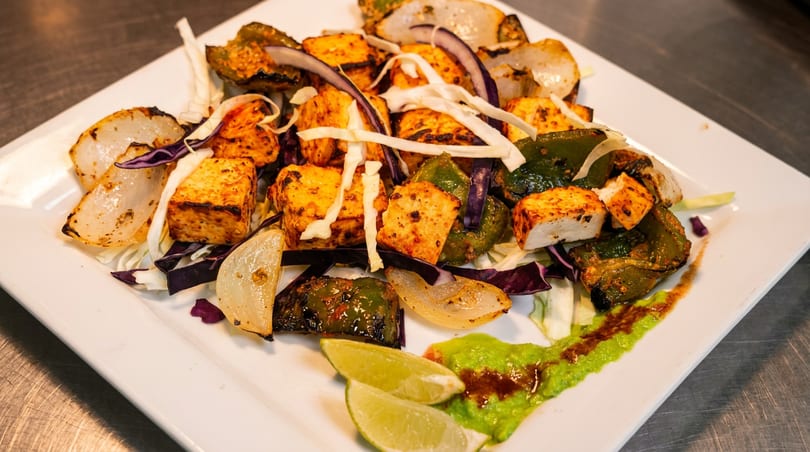 Grilled paneer, bell peppers, and onions with green sauce and lime wedges on a white plate.