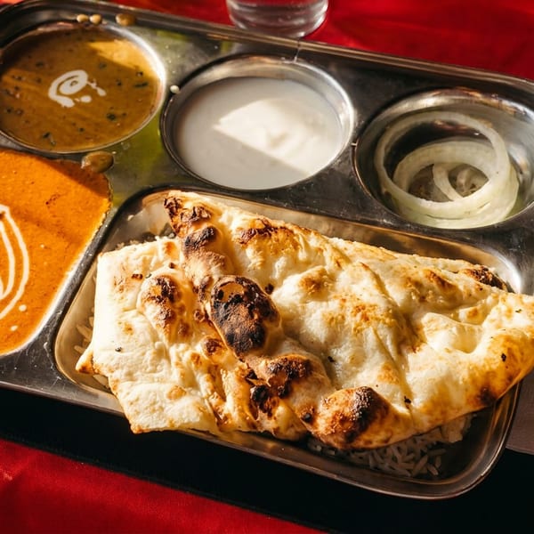 Indian thali with butter chicken, dal, yogurt, onions, rice, and naan bread on a red table.