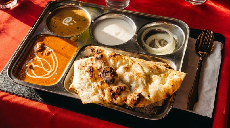 Indian thali with butter chicken, dal, yogurt, onions, rice, and naan bread on a red table.